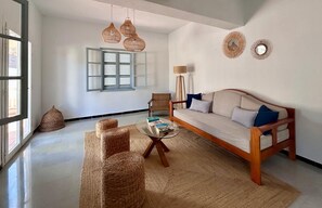Una sala de estar con un sofá, dos sillones, una mesa de centro, otomanas de mimbre, una lámpara de pie y una ventana con contraventanas.