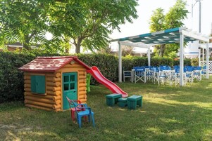 A log cabin playhouse with a red slide, surrounded by a grassy area and blue chairs.