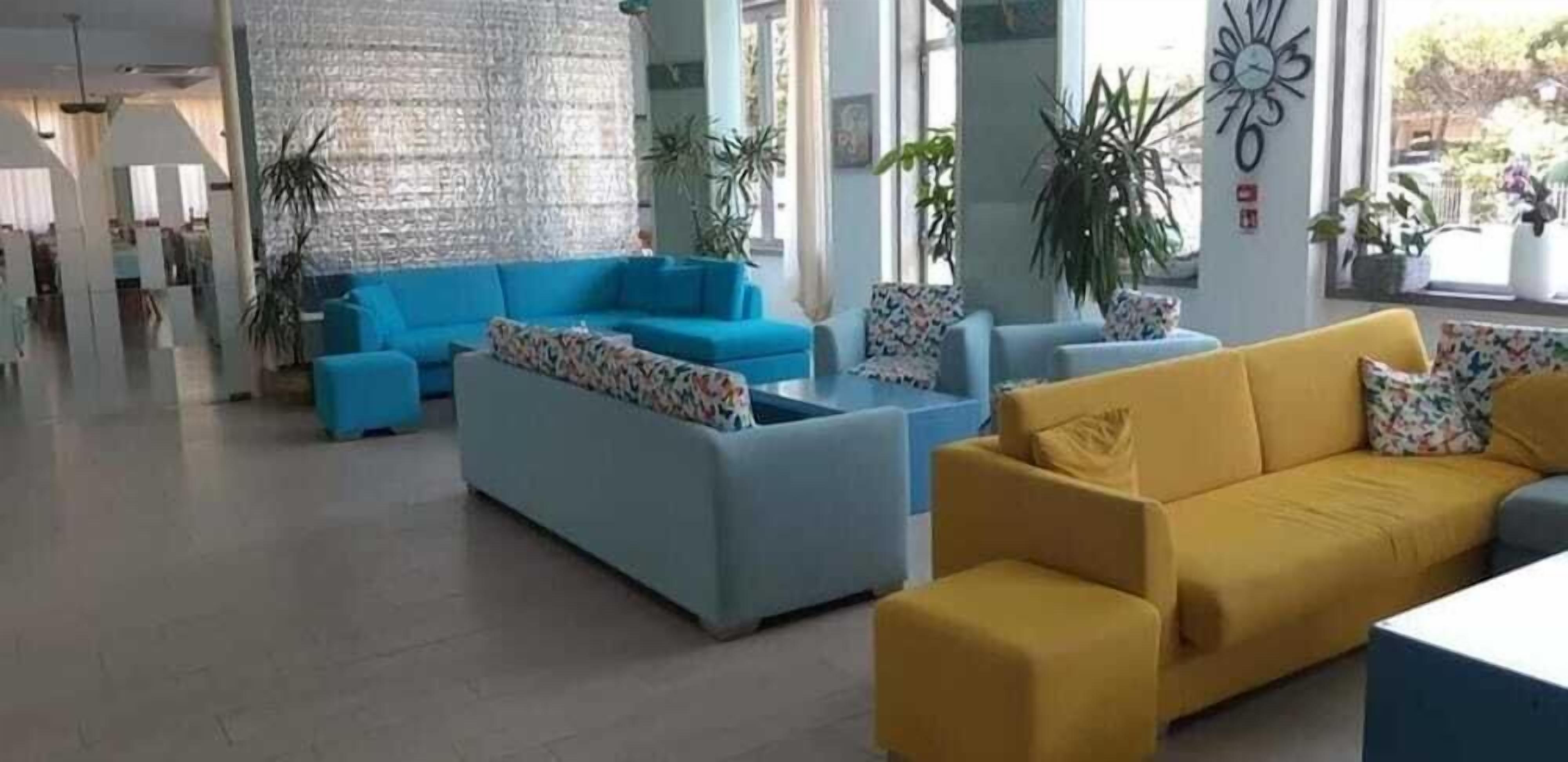 En moderne lobby med en blå sofa, en gul sofa og en grå sofa. Der er plantede planter og et stort vægur.