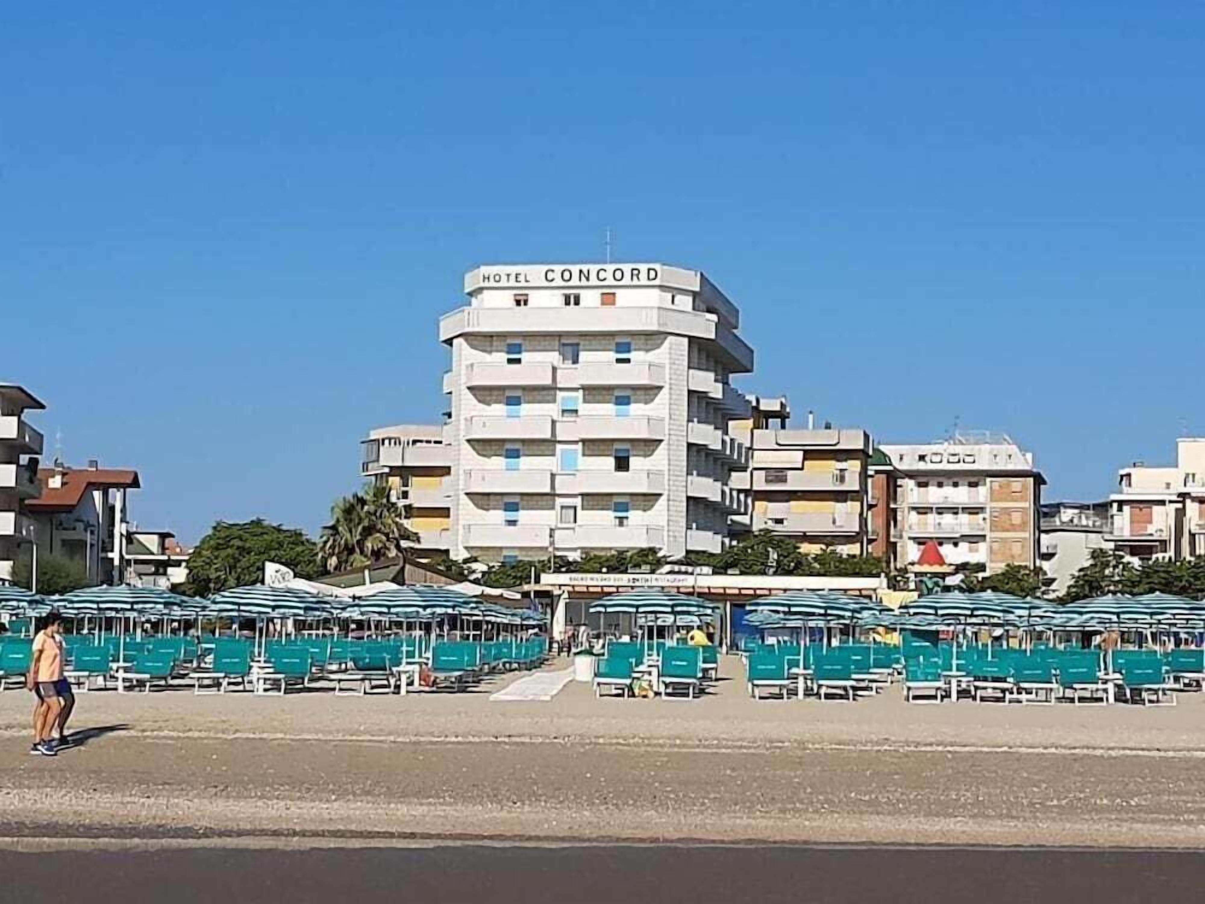 A hotel named 'HOTEL CONCORD' with multiple floors and balconies, a beach area with umbrellas and lounge chairs, and a clear blue sky.