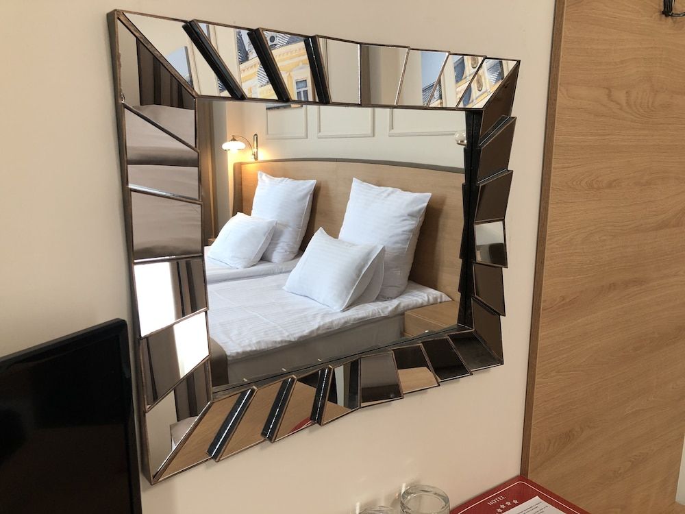 Una cama con ropa de cama y almohadas blancas, una mesita de noche con lámpara, y un espejo en la pared que refleja una escalera.