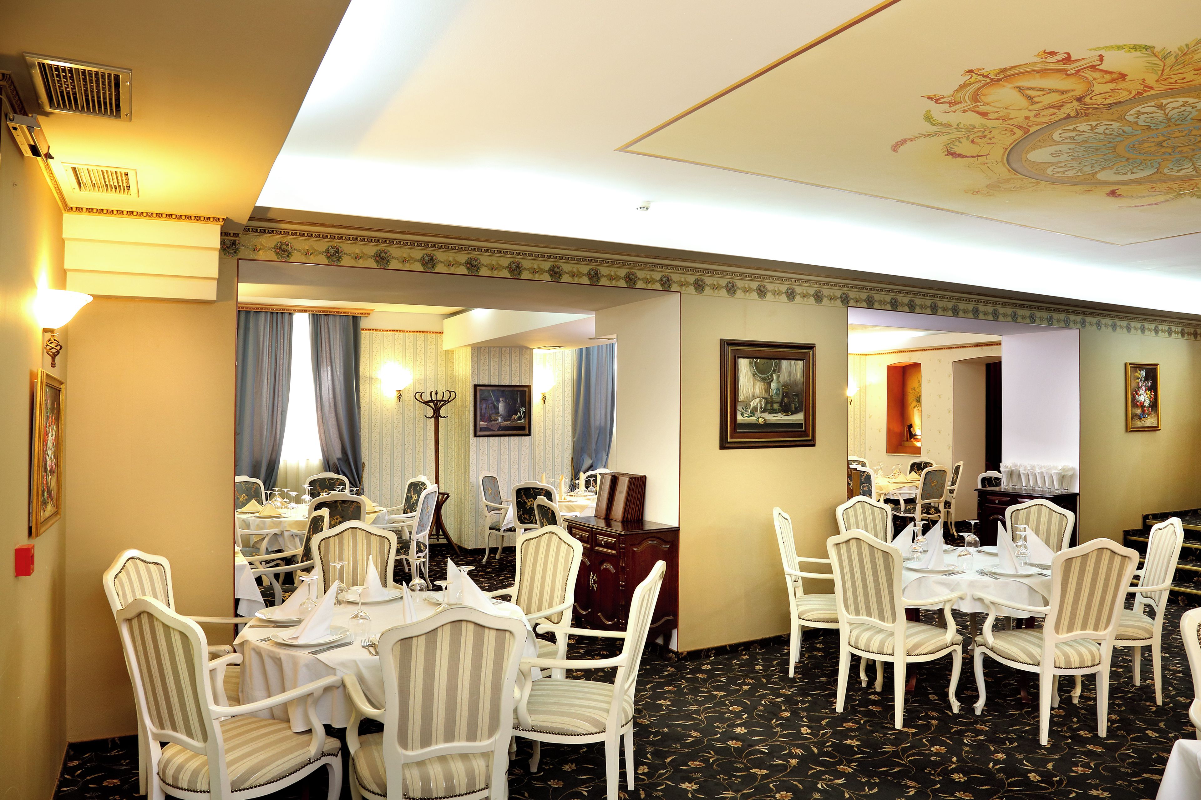 Ein Hotelrestaurant mit mehreren Tischen, Stühlen, einer Kristallleuchter und einer verzierten Decke.