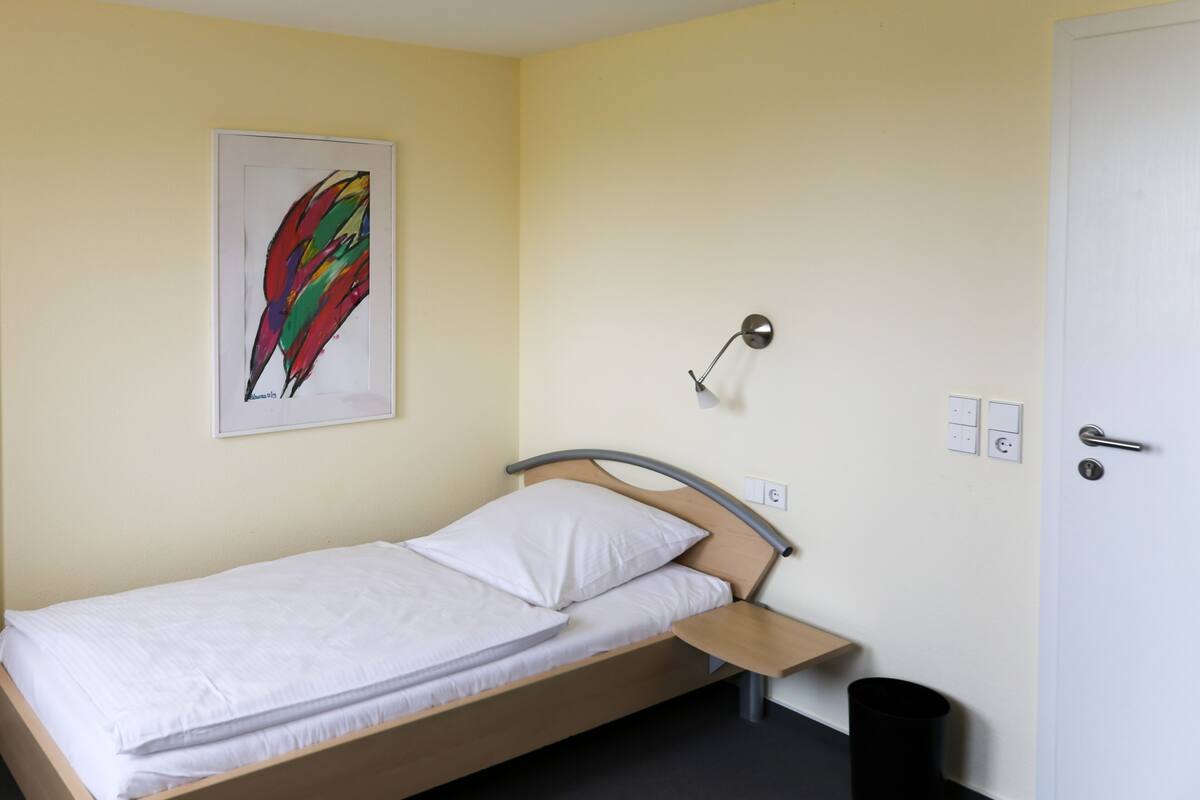 Een enkel bed met houten frame en een wit matras in een kamer met een lichtgele muur.