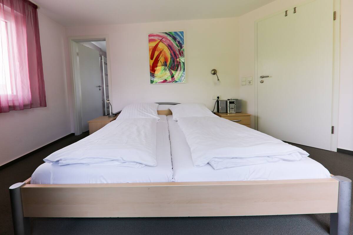 Een hotelkamer met een tweepersoonsbed, een nachtkastje met een lamp, een kleurrijk abstract schilderij aan de muur en een deur met een handgreep.