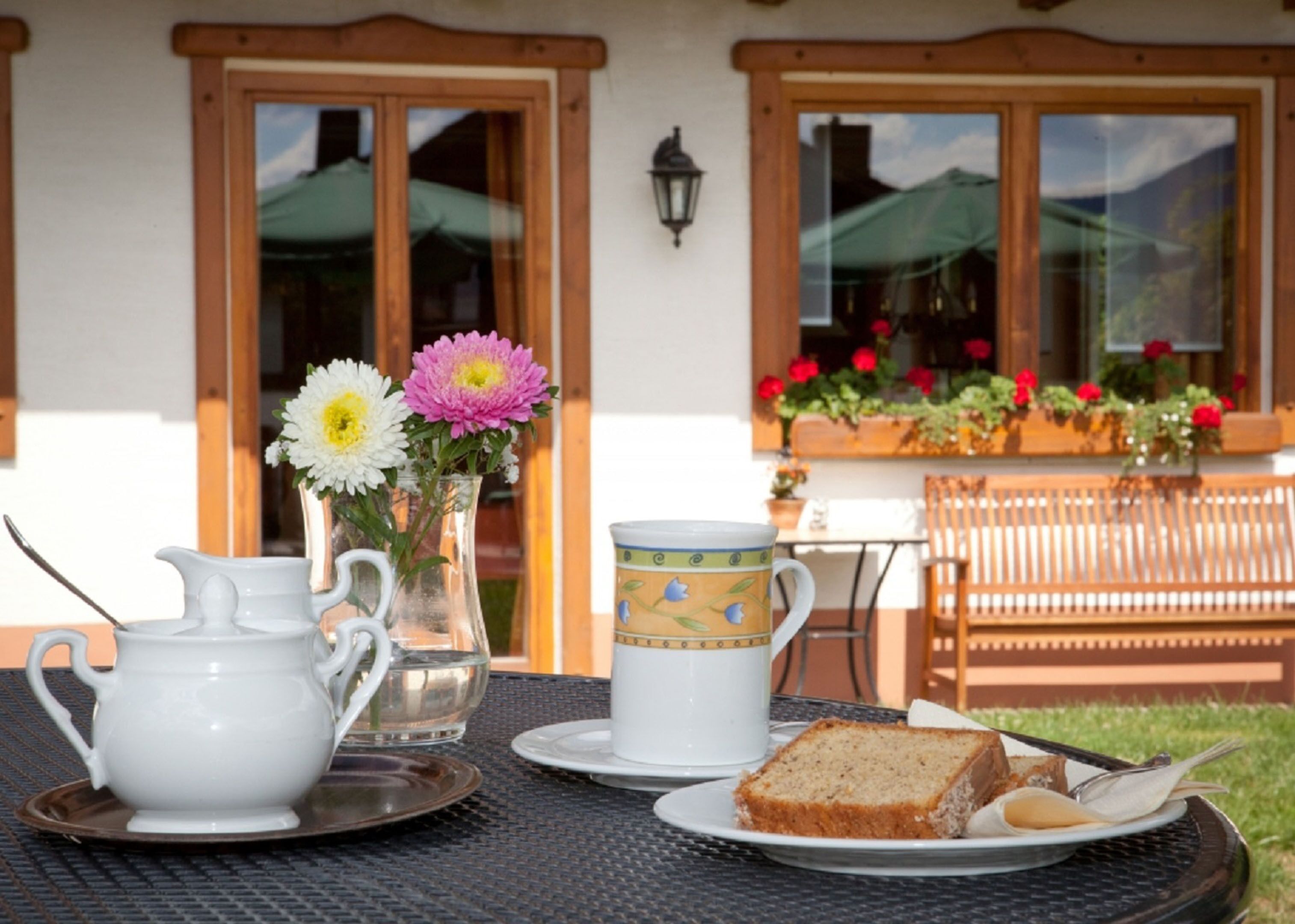 A table set with a teapot, jug, mug, and a slice of bread on a round table outside a building with wooden doors and windows.