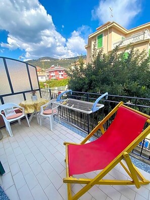 Un balcone con una sedia rossa, un tavolo e sedie, e vista su edifici e alberi.