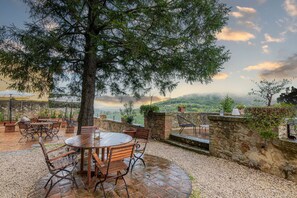 An outdoor patio area with round wooden tables and metal chairs, surrounded by stone walls and a large tree providing shade. - Palazzo Brandano (Trequanda)