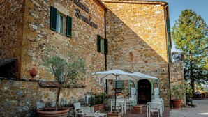 A stone building with a sign reading "Palazzo Brandino". There are outdoor tables and chairs, potted plants, and a large umbrella. - Palazzo Brandano (Trequanda)