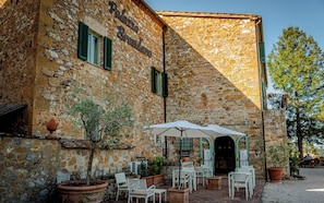 A stone building with a sign reading "Palazzo Brandino". There are outdoor tables and chairs, potted plants, and a large umbrella. - Palazzo Brandano (Trequanda)