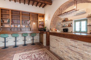 Un coin bar avec des étagères en bois, des livres et un mur en briques. On y trouve trois tabourets de bar verts et un tapis à motifs sur le sol.