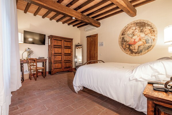 A bedroom with a bed, wooden ceiling beams, a television, a desk with a chair, and a wall-mounted painting.