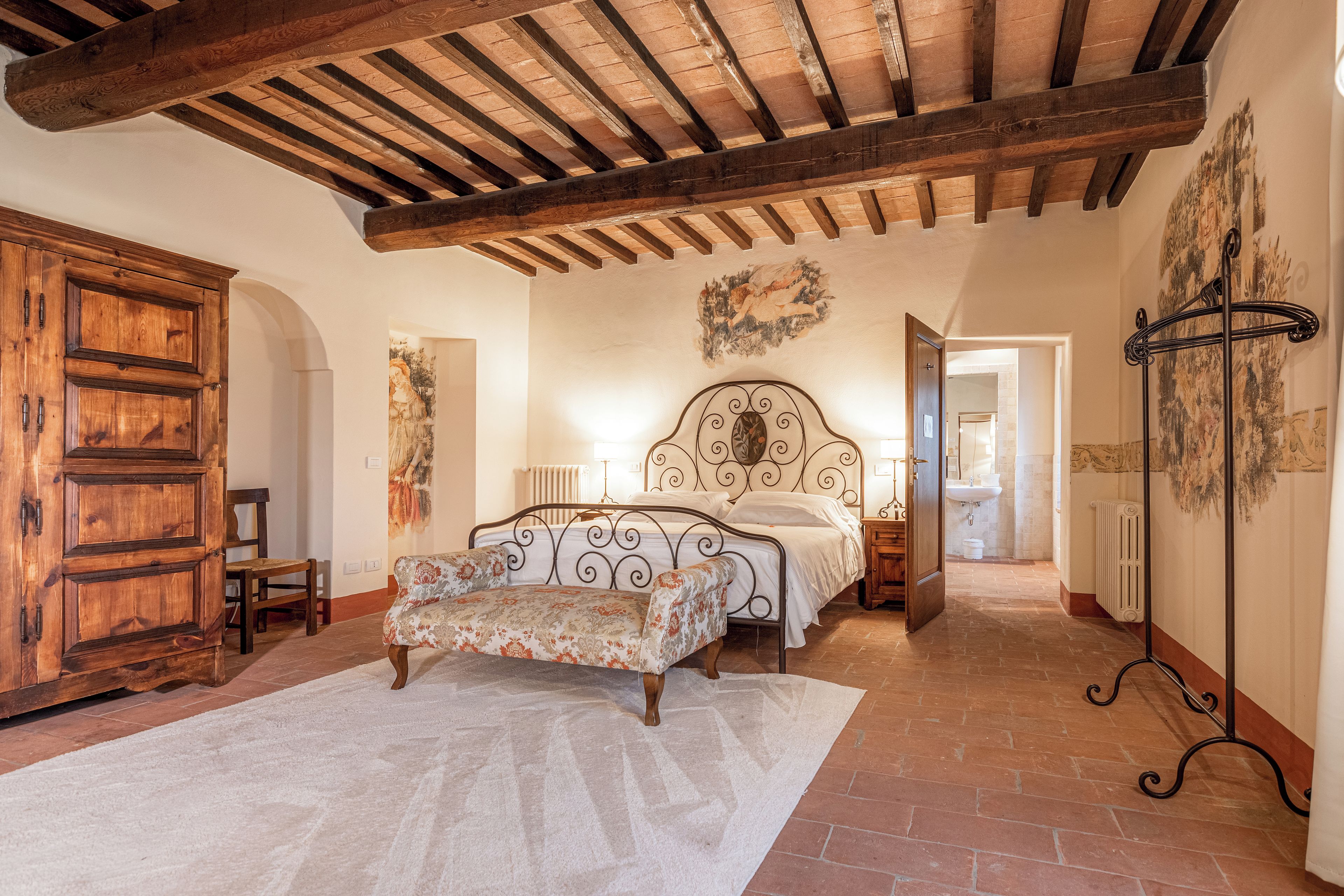A bedroom with a wooden ceiling, a bed with a decorative headboard, a bench, and a large wardrobe.