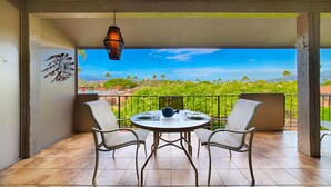 Un balcon avec une table ronde et deux chaises, une lanterne suspendue, et une vue sur un paysage verdoyant parsemé de palmiers.