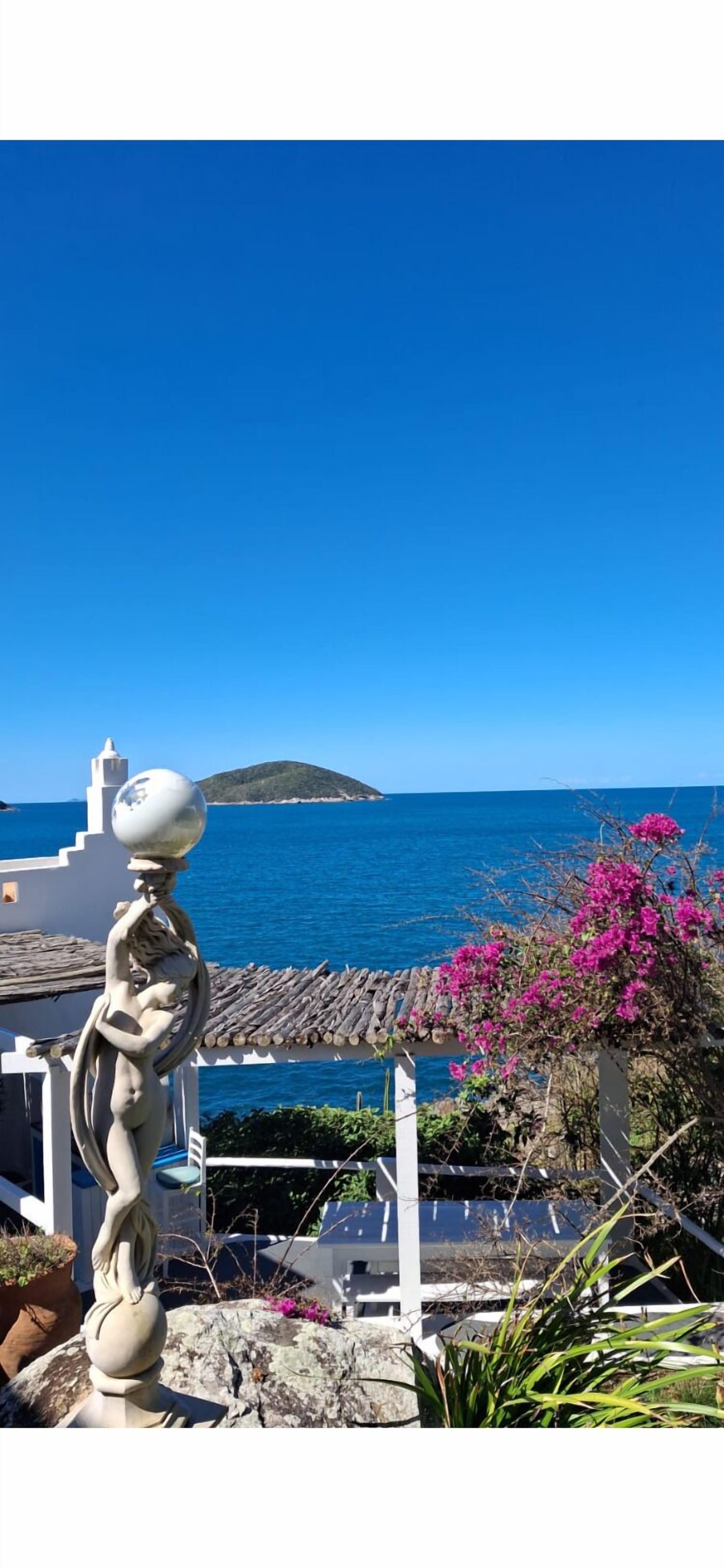A coastal view with a statue, a white building, and a pink flowering bush.