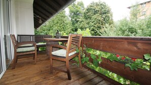 A wooden deck with two chairs and a table, a wooden railing with greenery, and a view of trees and buildings in the background. - Exclusive vacation in the historic building in Chiemgau   (Traunstein)