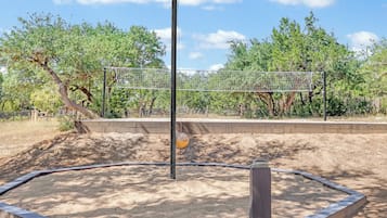 Un área de juegos con arena, aro y red de baloncesto, rodeada de árboles y una valla.