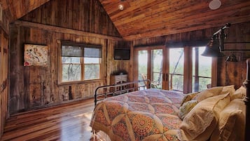A rustic wooden bedroom with a large bed, a television, and multiple windows offering a view of the outdoors.