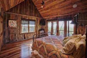 Quarto rústico de madeira com uma cama grande, televisão e várias janelas com vista para o exterior.