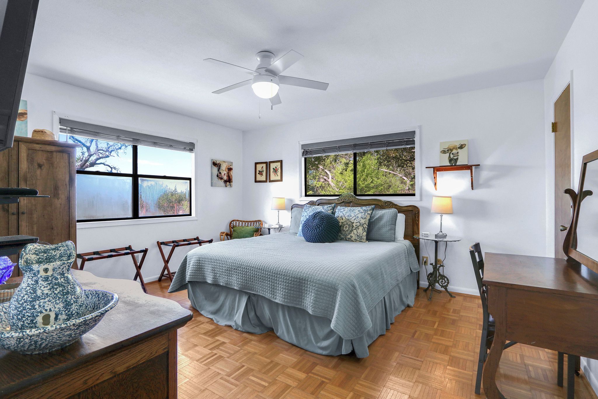 A bedroom with a bed, a ceiling fan, a window, a lamp, and a wooden table.