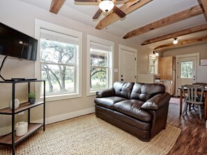 A living room with a leather sofa, a TV on a stand, and large windows. - Morning Dove I Romantic, Minutes from The Square! (Wimberley)
