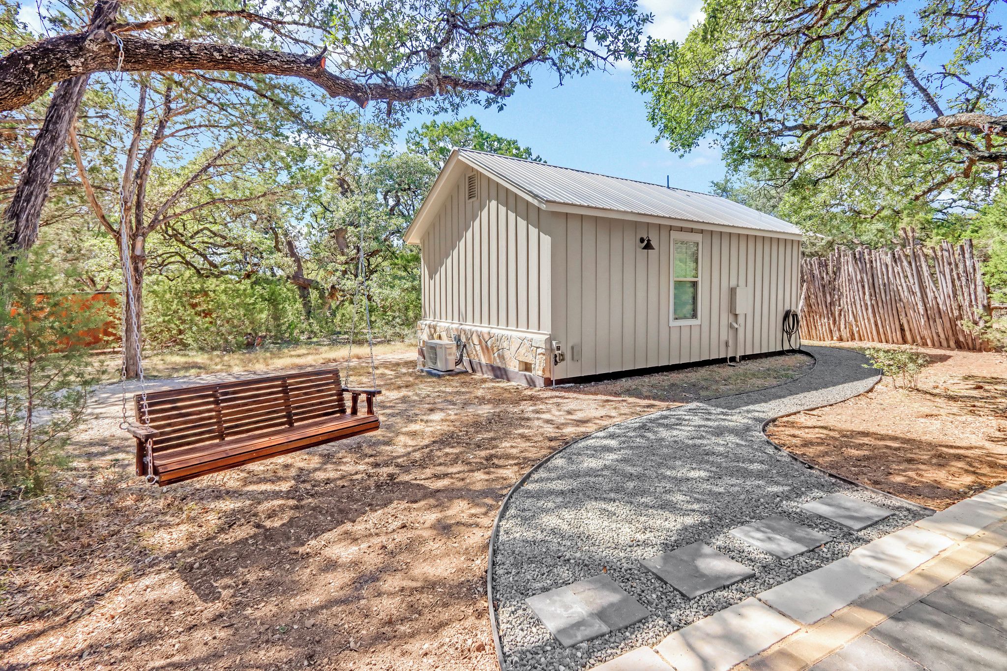 A small wooden cabin with a porch, surrounded by trees and a gravel path.