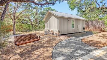 A small wooden cabin with a porch, surrounded by trees and a gravel path.