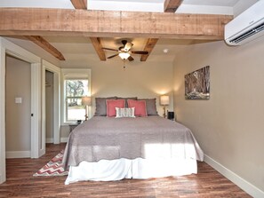 A bedroom with a large bed, wooden beams, a ceiling fan, and a window. - Morning Dove I Romantic, Minutes from The Square! (Wimberley)