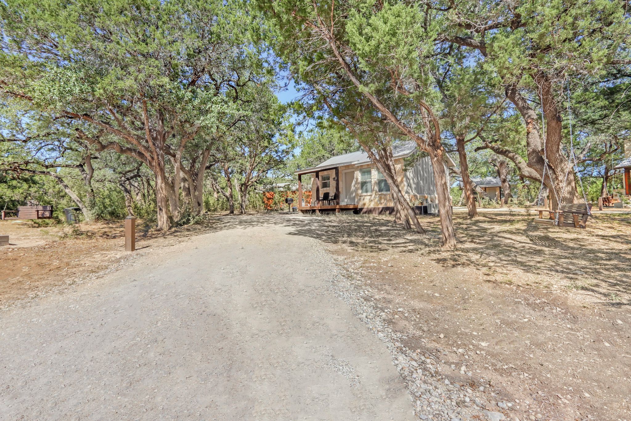A rustic cabin with a porch, surrounded by tall trees and a dirt road.
