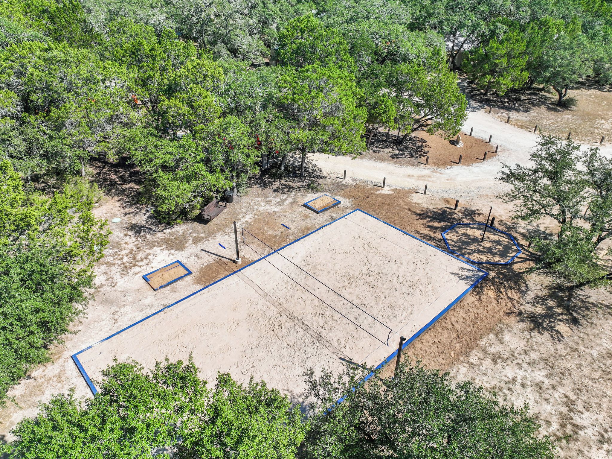 An aerial view of a sandy sports court with blue lines, surrounded by dense green trees.