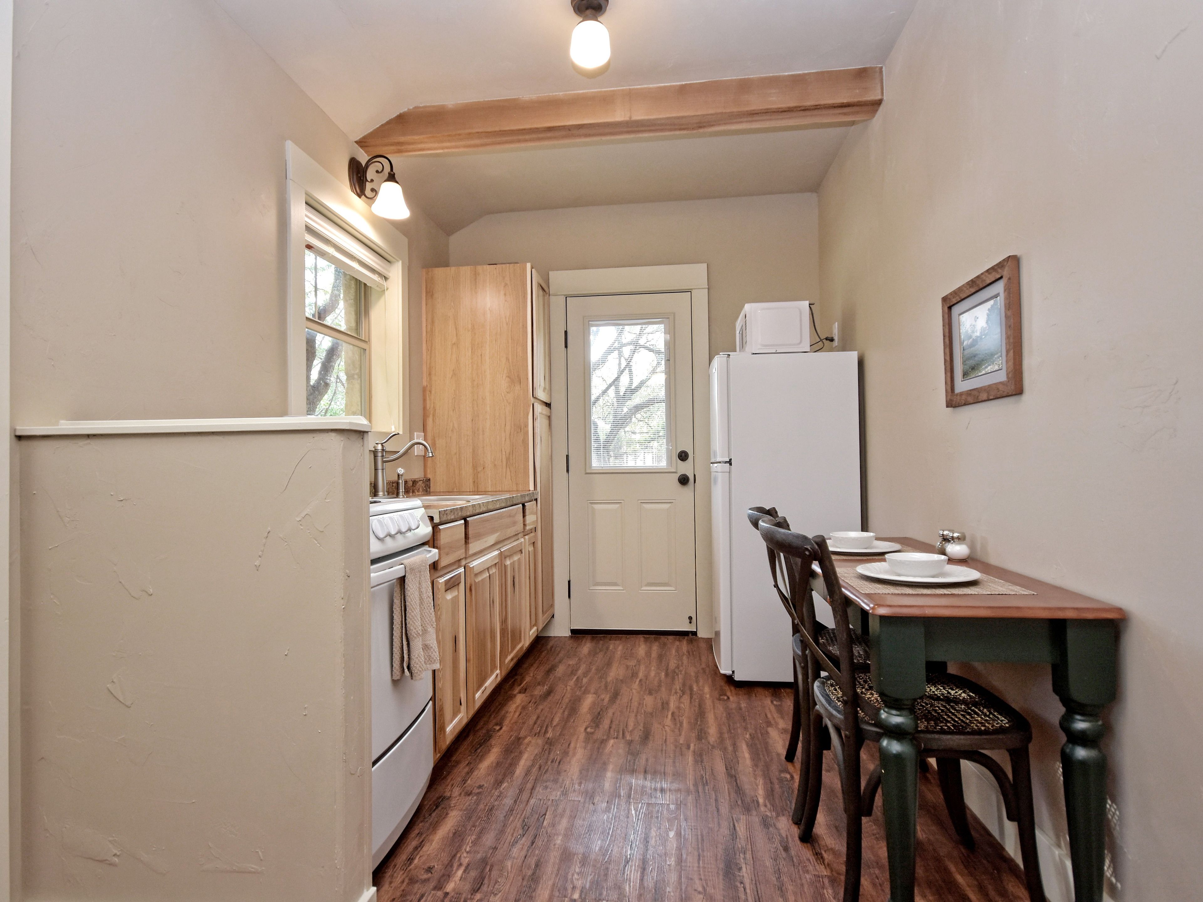 Una cucina compatta con mobili in legno, un frigorifero bianco, un tavolo con sedie e una finestra che si affaccia sugli alberi.