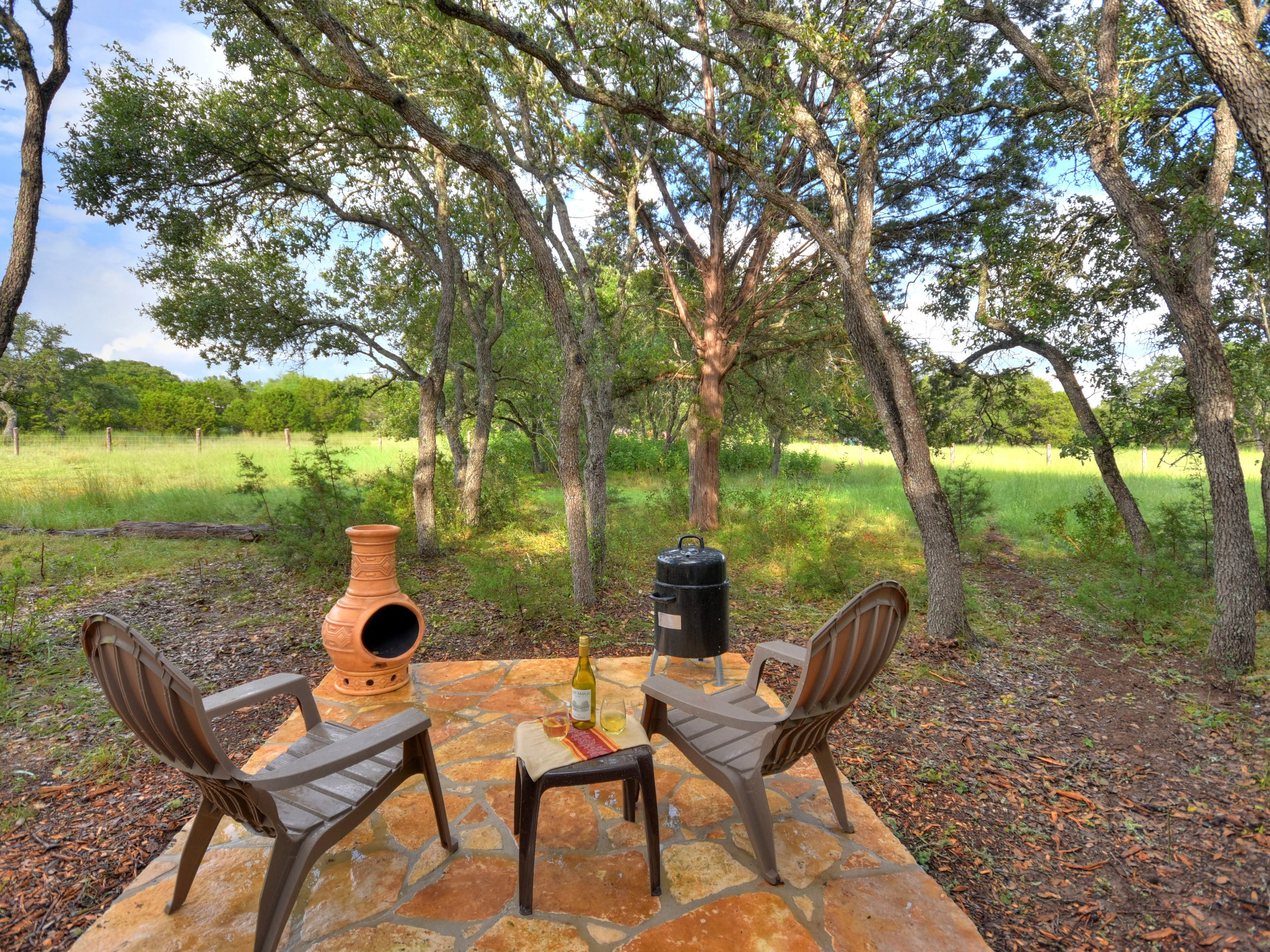 An outdoor seating area with two chairs and a table, surrounded by trees and a grassy field.