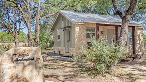 A rustic cabin with a stone facade and a metal roof, surrounded by trees and a clear sky. - HCPL-Mockingbird I Cabin, Close to Wimbeley Square! (Wimberley)