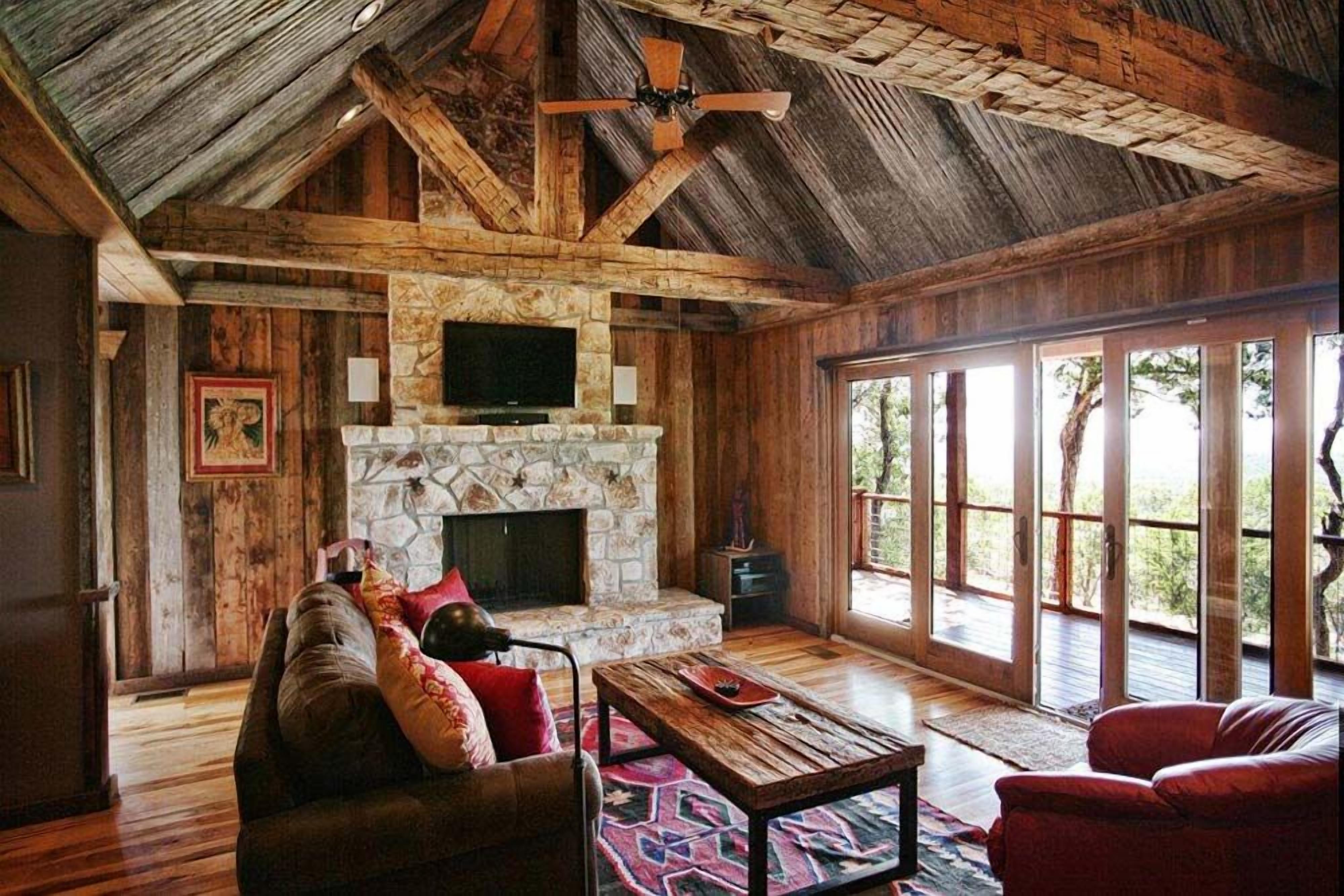 A rustic interior with a stone fireplace, wooden beams, a ceiling fan, and large windows.