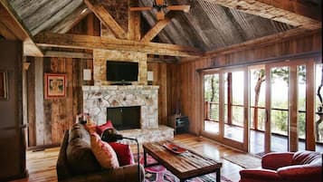 A rustic interior with a stone fireplace, wooden beams, a ceiling fan, and large windows.