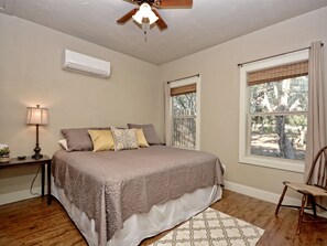 A bedroom with a bed, a nightstand, a lamp, a chair, a window with a view of trees, and a ceiling fan.
