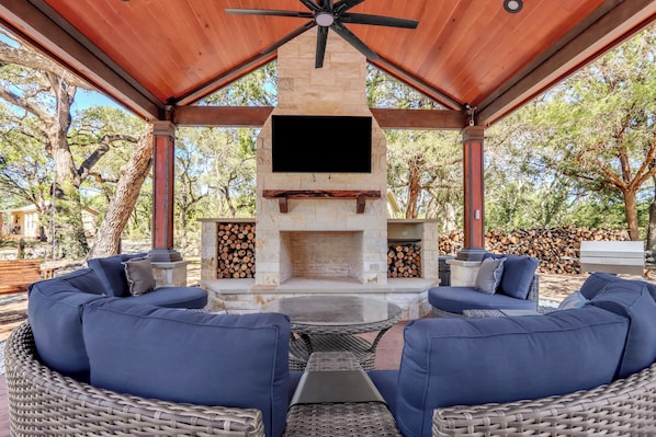 An outdoor patio with a stone fireplace, blue cushioned seating, and a flat-screen TV mounted above the hearth.