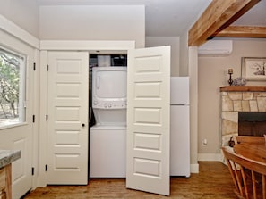 A kitchen with a built-in washer and dryer, a stone fireplace, and a dining area with a wooden table and chairs. - Mountain Laurel I 2Br Cabin near Square! (Wimberley)