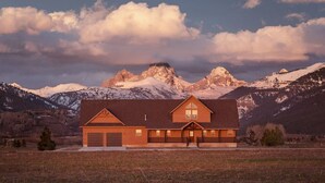 Eine zweistöckige Holzhütte mit Garage, vor einer Kulisse aus schneebedeckten Bergen und einem bewölkten Himmel.