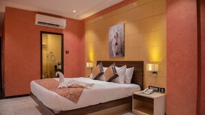A hotel room with a large bed, a wooden headboard, a painting on the wall, a bedside table with a lamp, and an air conditioning unit.