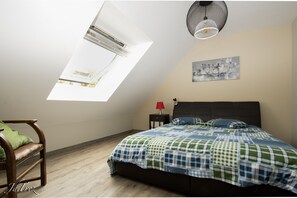 A bedroom with a sloped ceiling, a bed with a checkered bedspread, a nightstand with a red lamp, a chair with a green cushion, and a wall-mounted artwork.