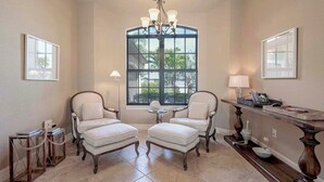 Une pièce avec deux fauteuils blancs, un lustre, une grande fenêtre donnant sur un beau paysage et une console en bois surmontée d’une lampe.