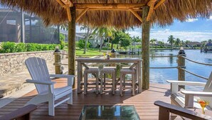 Une terrasse en bois avec des chaises blanches et une table, donnant sur une étendue d’eau et surmontée d’un toit de chaume.