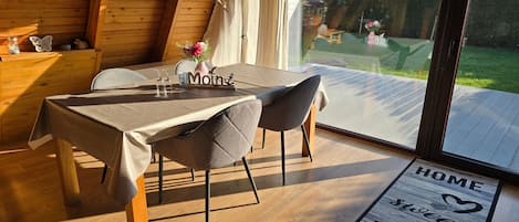 A dining area with a table set for two, a wooden interior, and a view of the garden through large windows.