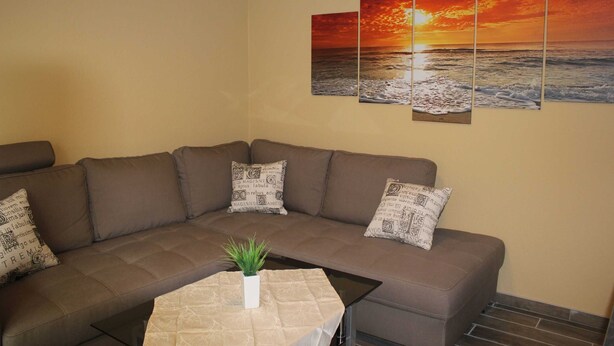 A living room with a brown sectional sofa, a small table with a plant, and a wall with a sunset painting.
