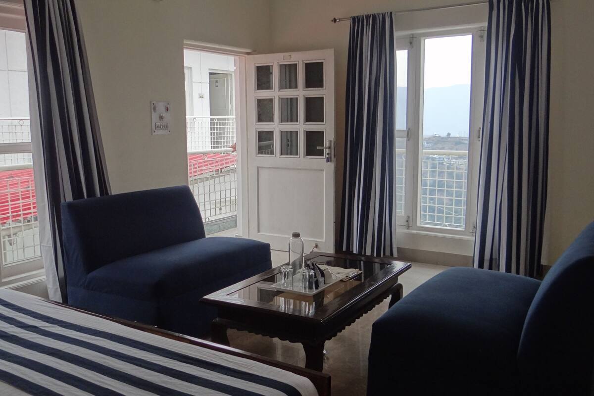 A hotel room with a bed, two armchairs, a small table with a bottle and glasses, and a view of a red building through a window.