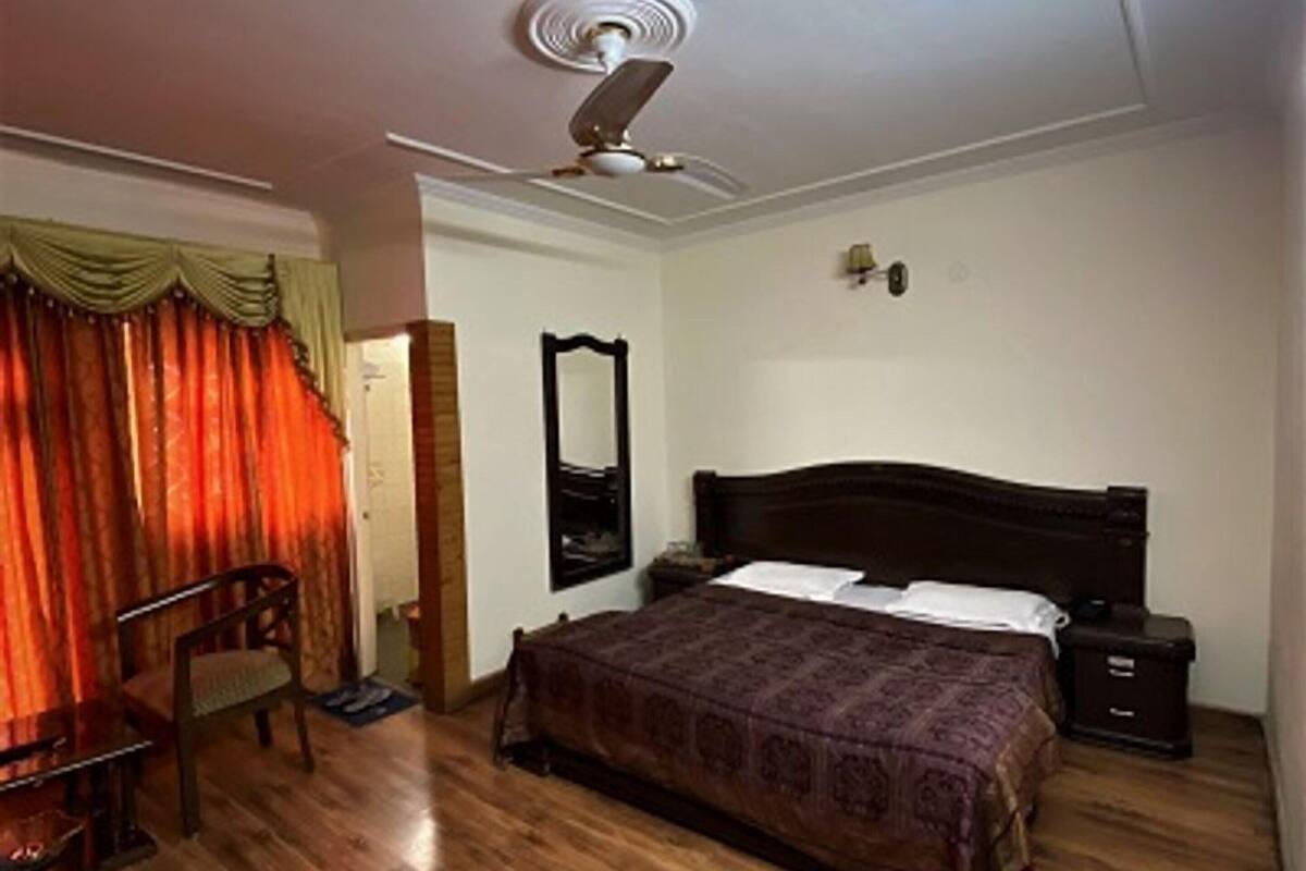 A bedroom with a wooden floor, a bed with a dark headboard, a chair, a bedside table, a mirror, and a ceiling fan.