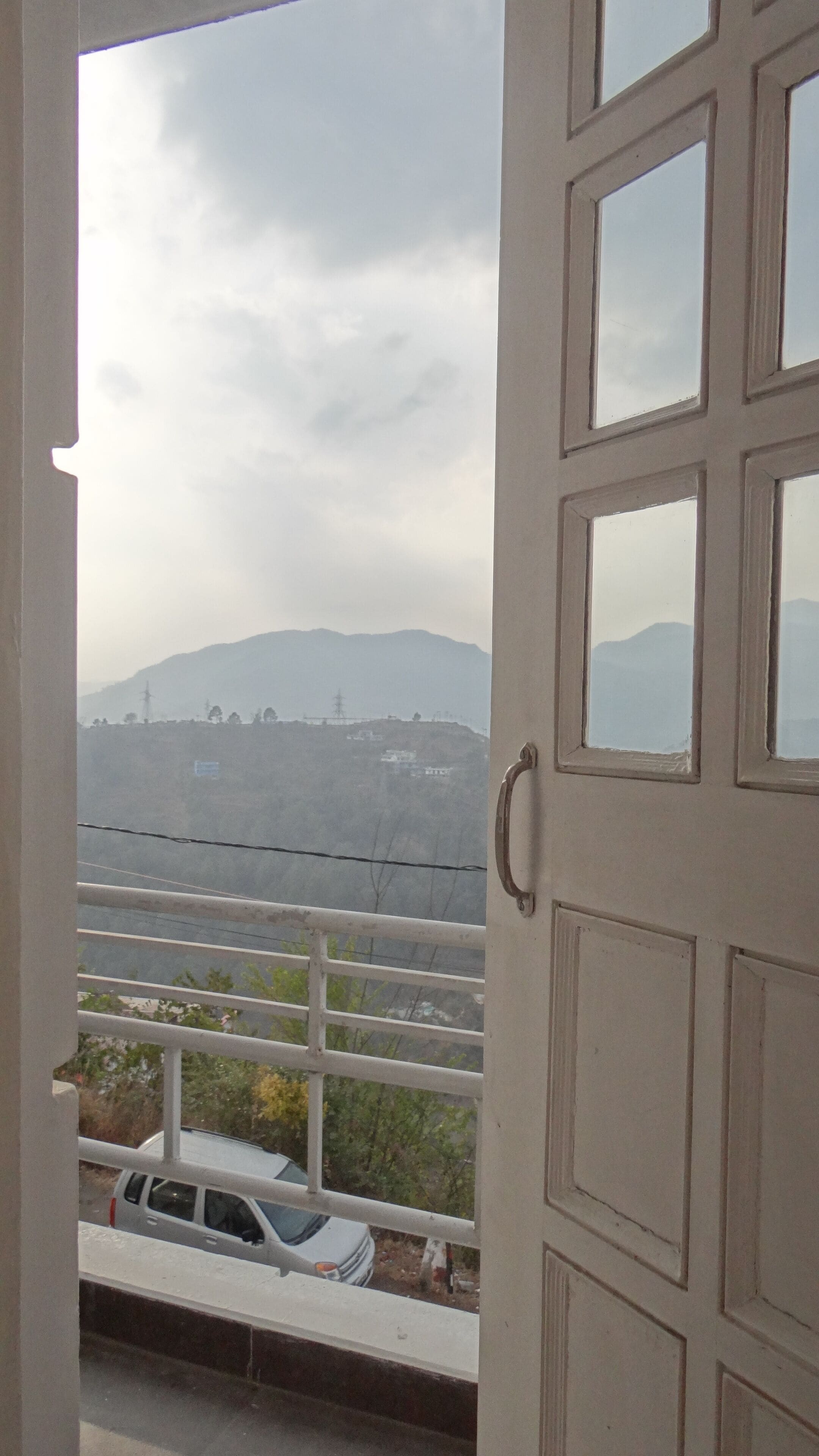 A view from a window with a metal railing, overlooking a road with parked cars and distant mountains.