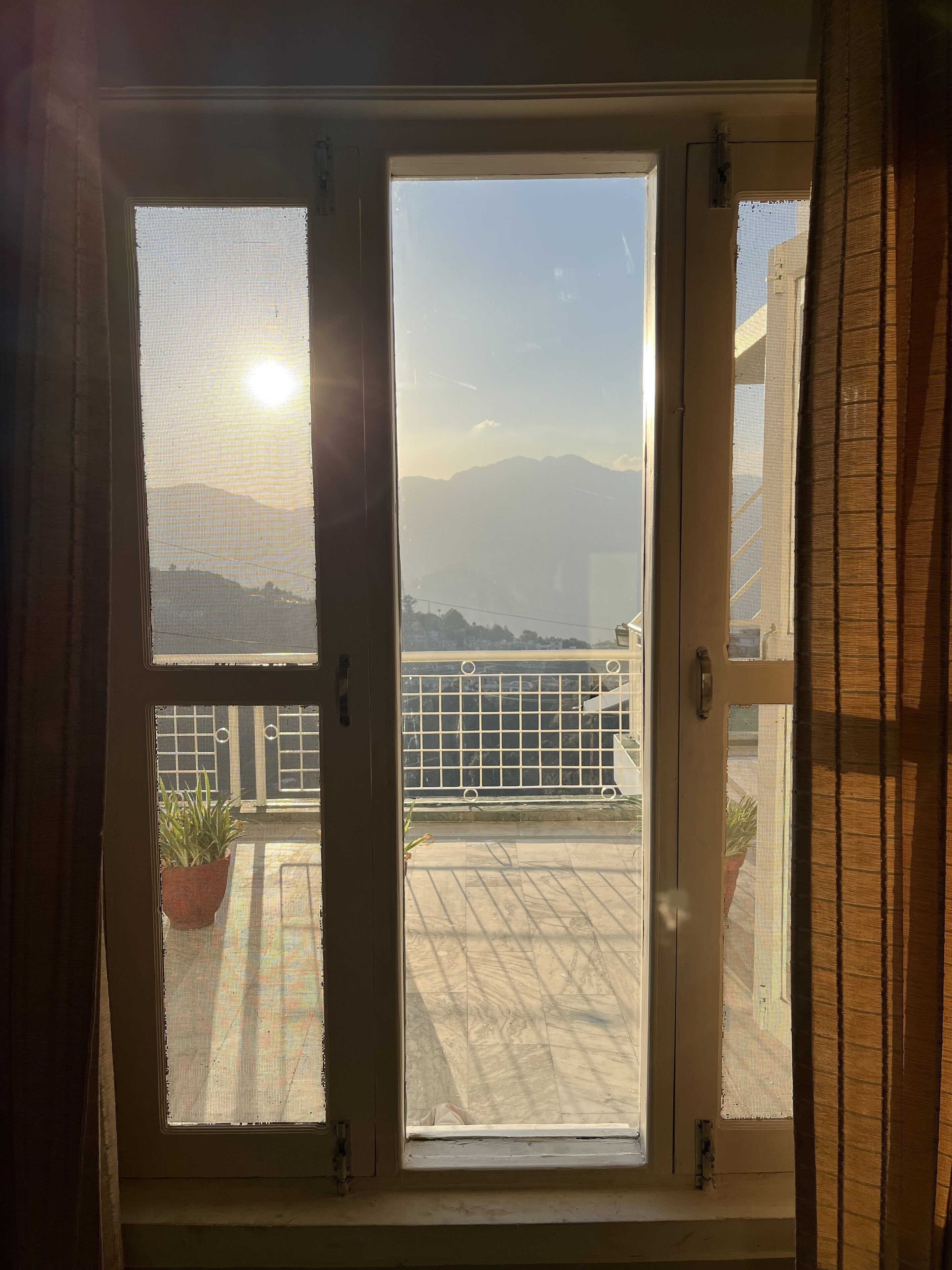 A view through a window with a view of a balcony, a railing, and a distant mountain range.