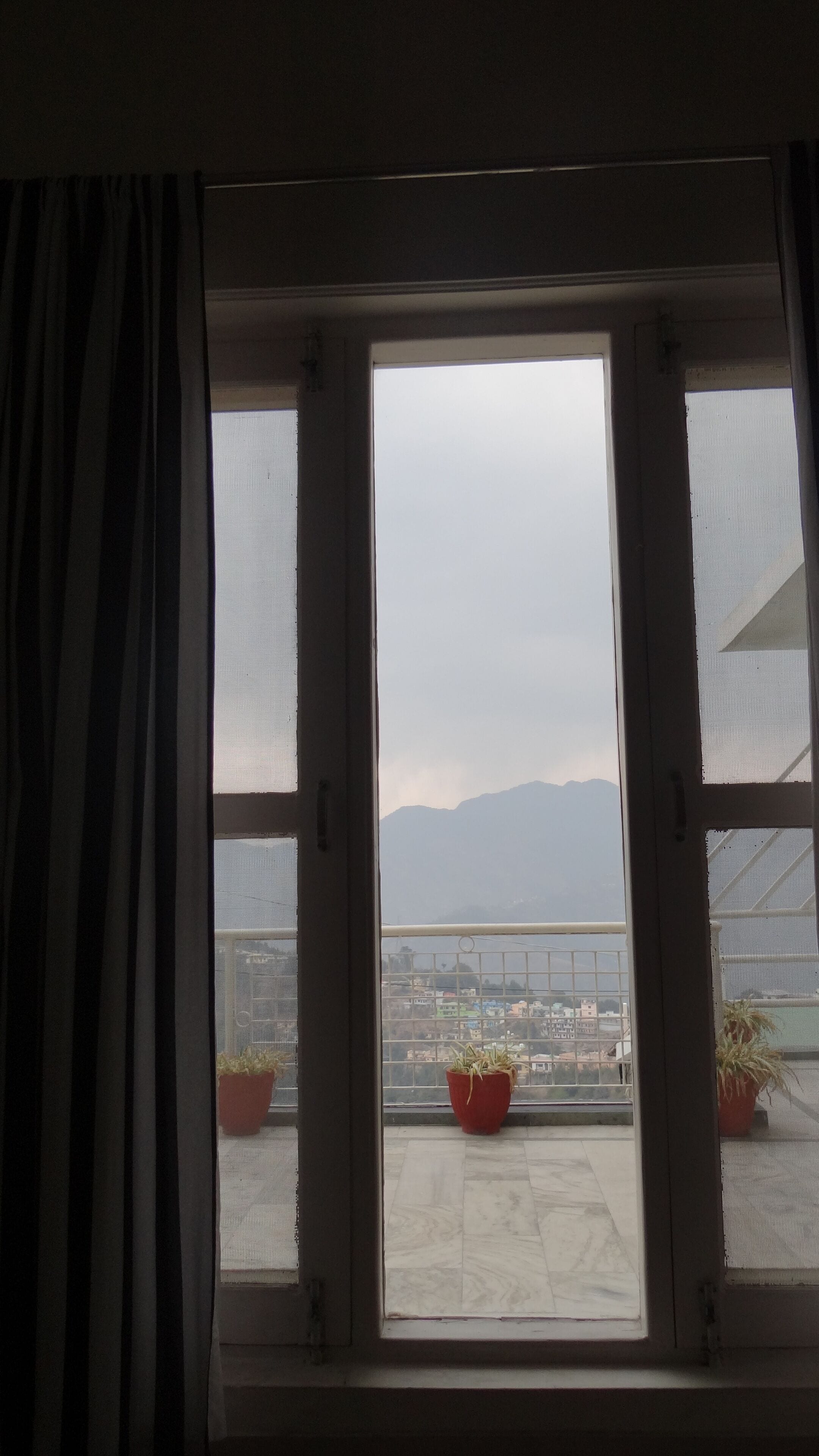 A window with a view of a balcony, potted plants, and distant mountains.