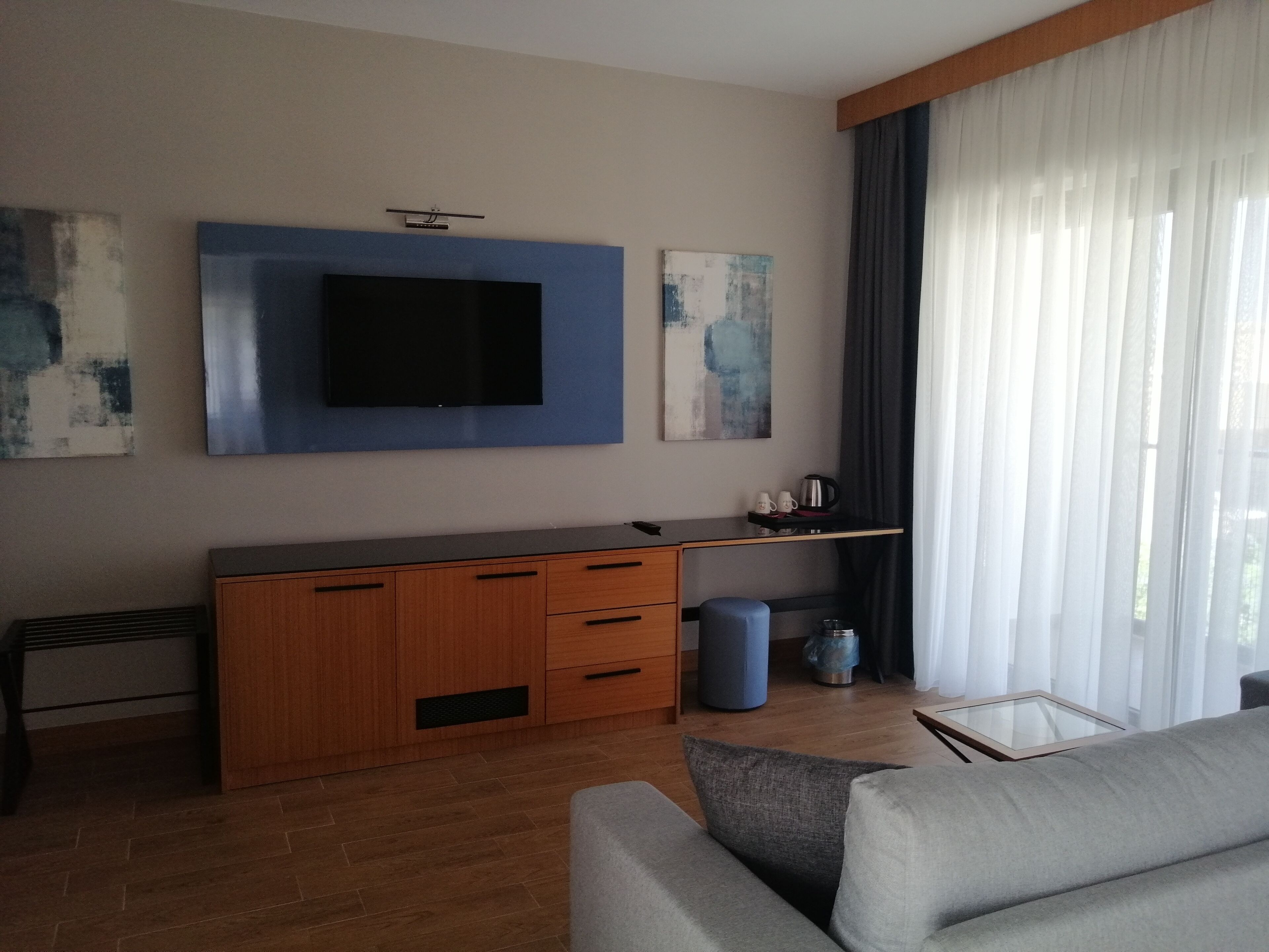 Un salón moderno con un televisor de pantalla plana montado en una pared azul decorativa, muebles de madera, una mesa de centro de cristal y un ventanal con cortinas translúcidas.